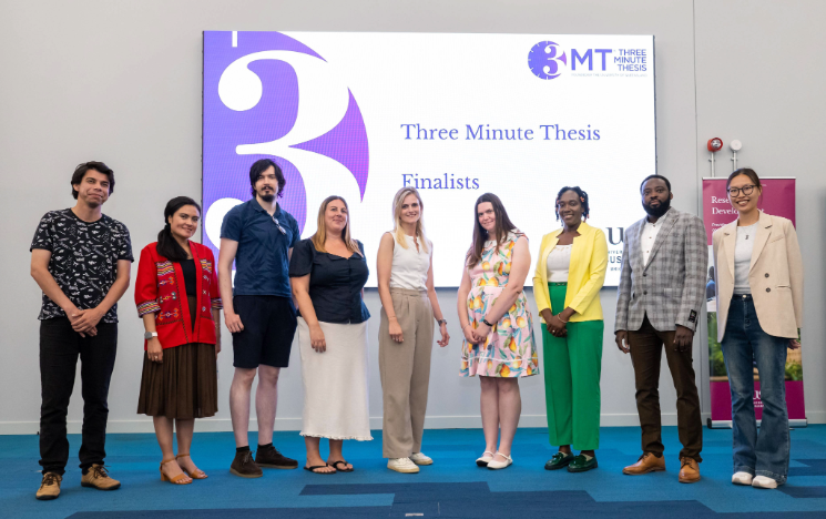 A group of nine people line up and smile on stage in front of a Three Minute Thesis Finalists presentation screen
