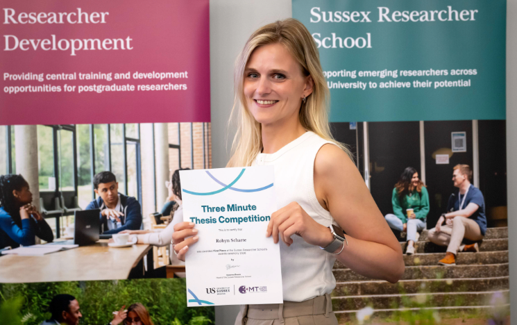 A Three Minute Thesis Competition winner shows their first-place certificate with two Sussex Researcher School banners in the background