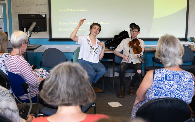 A presenter gestures with both hands while speaking and sitting next to a violinist, performing in front of a seated audience and a presentation screen.