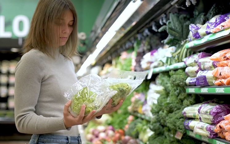 Woman shopping in a supermarket produce aisle, holding and looking at a bag of green lettuce beside shelves of fresh vegetables.