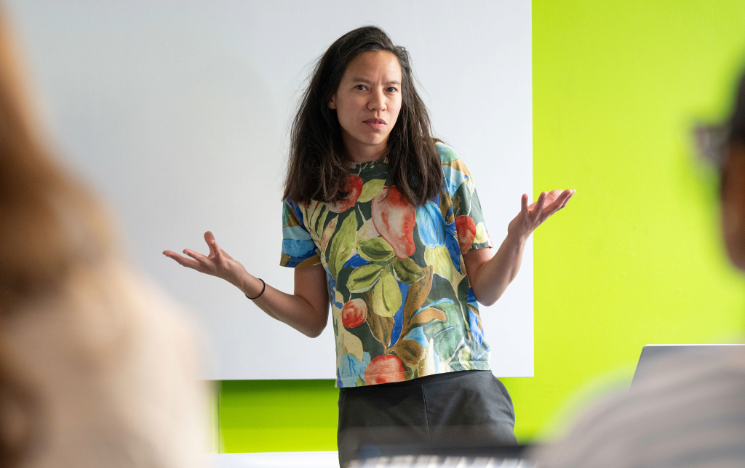 A presenter wearing a colorful fruit-patterned t-shirt gestures with open arms while presenting in a room with a lime green wall.