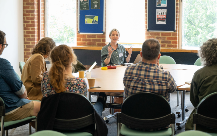 A workshop leader speaks and gestures with both hands while leading a discussion with a group seated around a circular table.
