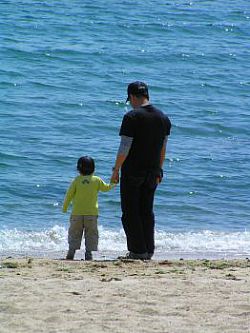 A photo of a father with his child, standing on a beach holding hands and looking out to sea