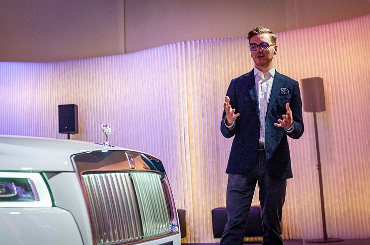 Photo of alum Rhodri Good in a suit presenting a new Rolls Royce, the bonnet of which is also in the photo.