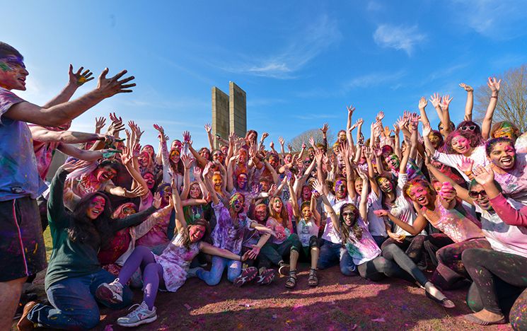 Large gathering of students, covered in coloured paint from Holi celebrations, seated and standing with arms in the air, in front of campus Arts buildings