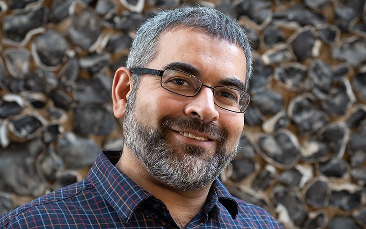 Professor Robin Banerjee, smiling at camera, portrait head and shoulders photo in front of campus flint wall