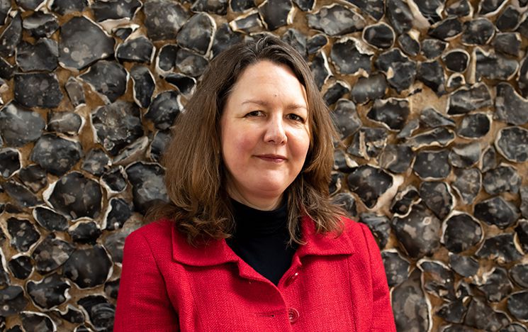 Professor Liz David-Barrett, smiling at the camera, head and shoulders portrait shot, in front of campus flint wall