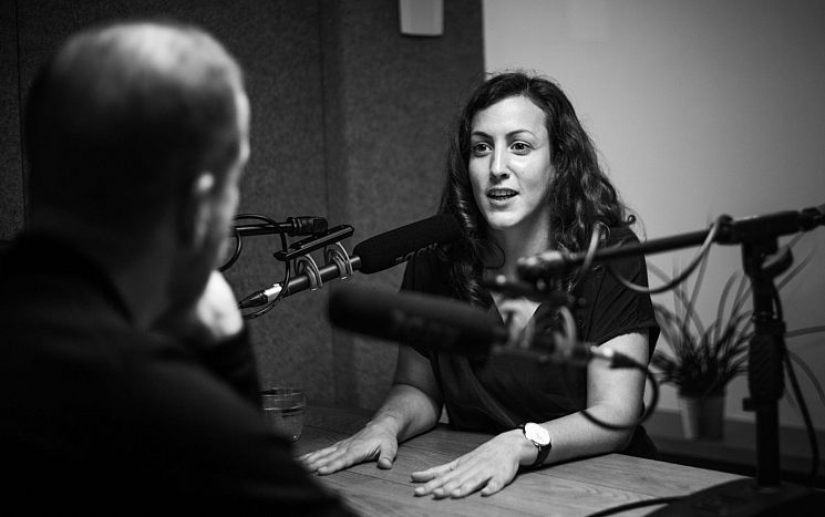 Black and white photo of alumna Sara Osterholzer seated at desk with microphones, recording podcast