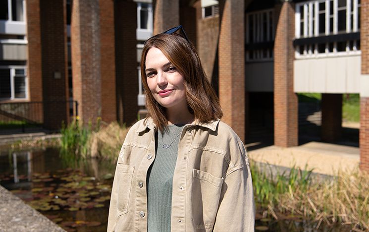 Louise Harman standing facing the camera on campus with Arts pond and buildings in the background