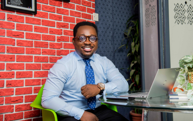 Alumnus Gideon sits at a table with a laptop. He is wearing a blue shirt and brown glasses.