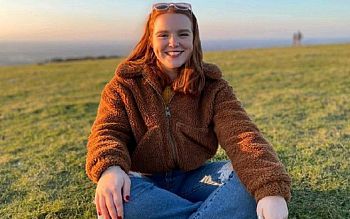 Photo of Zoe sitting on the grass, legs crossed, smiling at the camera, with sea and sky in the background