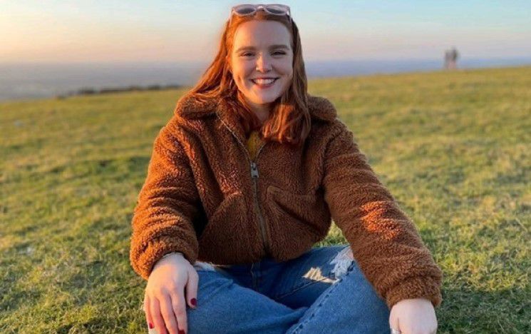 Alumna Zoe Bonnell seated on the grass, with legs crossed, smiling at the camera, with sea and sky background