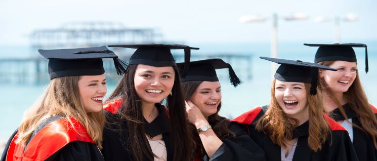 Five female graduating students in their caps and gowns on Brighton beach with the west pier in the distance