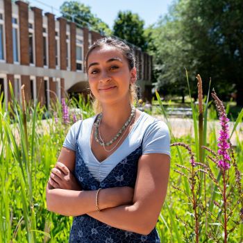Lottie Gomes, Arts Scholar, standing with flowers behind her and a campus building in the distance