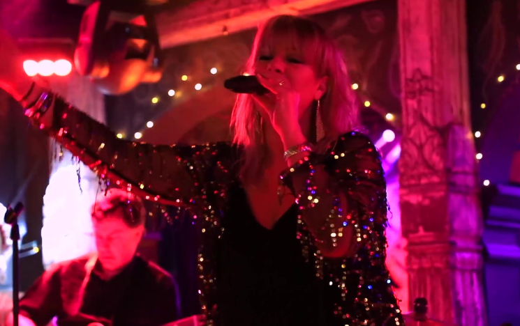 Helen Nagle singing, with microphone, as she performs with her band wearing sparkly top, guitarist in the background,