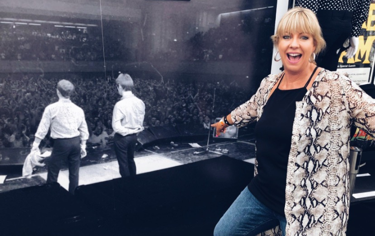 Helen visiting an exhibition in Brighton about The Jam, she stands smiling in front of a large black and white photograph of the Jam performing