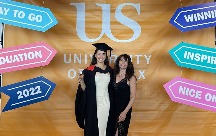Corinne Furman in her graduation gown with mum, and a university of sussex graduation graphics backdrop