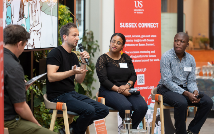 Omid Moallemi speaking to the crowd, as speakers Emnet Tiruneh and Benjy Kusi listen sat on stools beside him.