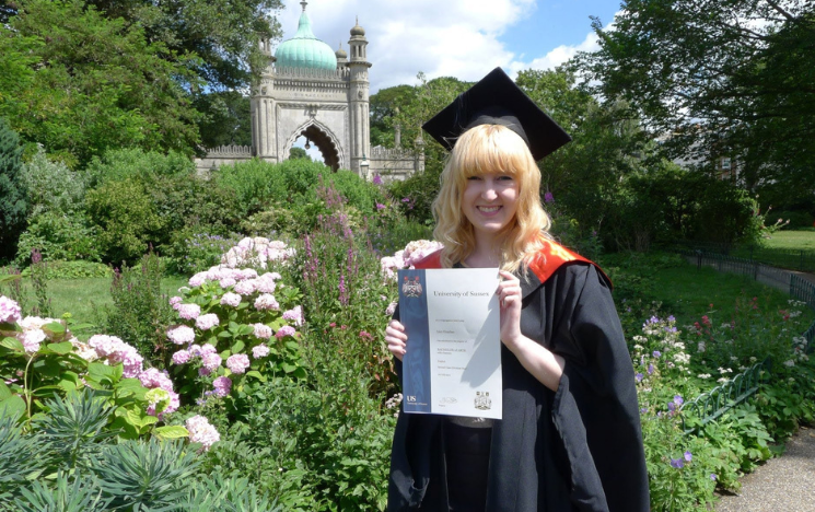 Lucy posing in the Royal Pavilion Gardens with her degree certificate. She is wearing a graduation gown and mortar board.