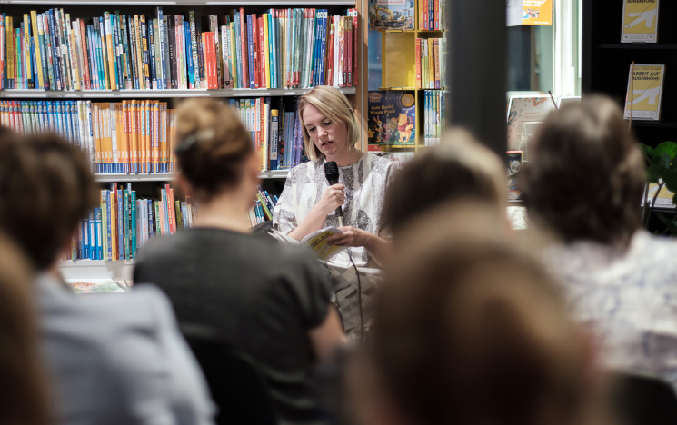 Lena Marie Glaser reading from her book in front of an audience sitting on chairs. There are lots of books behind Lena.