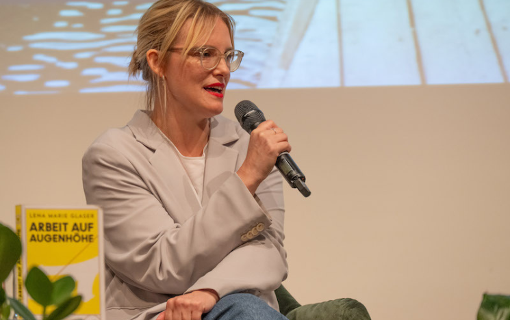 Lena Marie Glaser sat speaking at a panel with a microphone in her hand. Her book is stood up beside her.