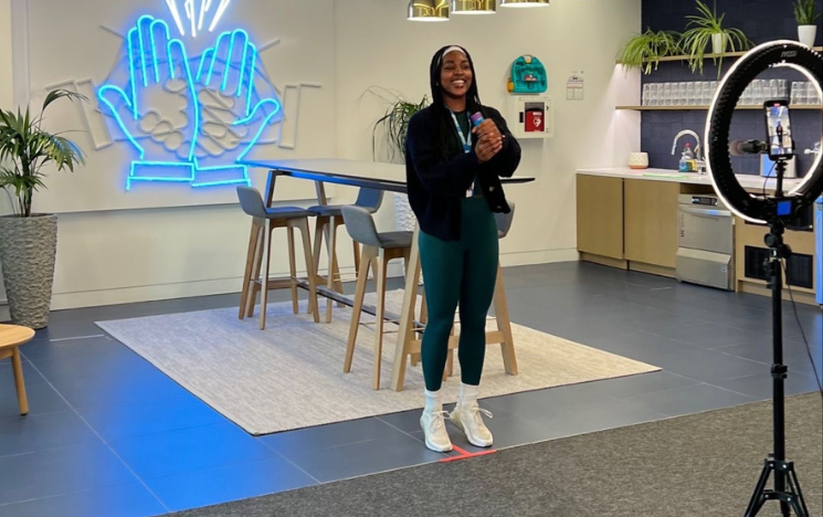 Jamila stood up filming in front of a ring light and camera. Behind Jamila is a neon sign of hands clapping.