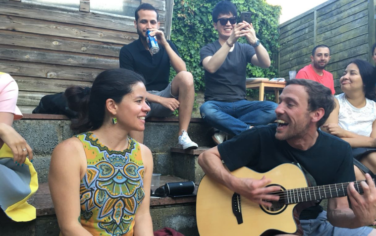 Jon McGlashan playing the guitar sitting next to Carolina Avellaneda on the grass. On the background, from left to right, a person sipping a beverage, another taking a picture and three other people smiling are sitting on the stairs.
