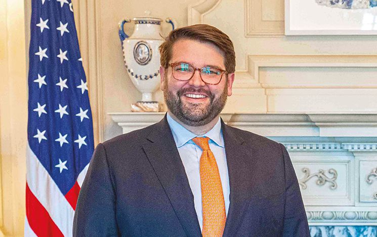 US diplomat Bill Russo in suit, smiling, with USA flag behind him