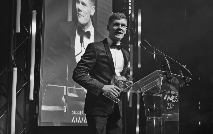 Black and white photograph of Olly Watts on stage in a suit receiving his Future Talent of the Year award at the Sussex Business Awards 2024.