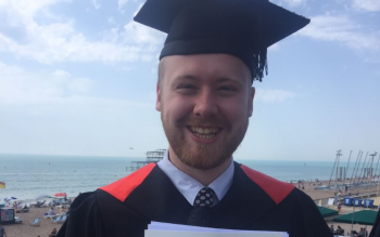 Shaun Ring in a graduation gown and mortar board posing in front of Brighton & Hove beach.