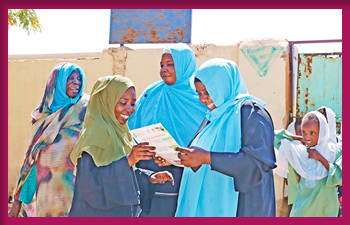 A group of women and a child, all wearing headscarves, smile and engage with a booklet outdoors near a worn wall and gate.
