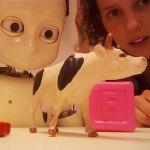 A robot looking at a toy cow and a toy block while Katherine Twomey looks on from behind.
