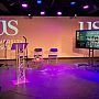 An empty but lit stage with a University of Sussex backdrop, a TV screen showing theme title of event, and podium