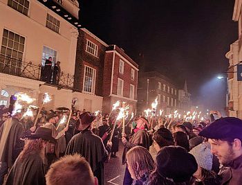 picture of lewes bonfire participants walking down a street holding fire lanterns. crowds line the street.