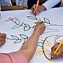 hands of four different people drawing leaves and flowers on a large white piece of paper with colored felt tip pens