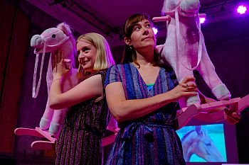 Two women wearing sparkly jumpsuits holding pink rocking horses. Photo of a horse on a projector behind them.