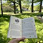 Hand holding a open children's book which has emoji stickers on it. Location is outside the Sussex Meeting House grassy area with trees.