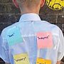 A child with short blond hair faces a brick wall. Four sticky notes on the back of their shirt read "Rude," "Disruptive," "Naughty," and "Lazy." A sad face is drawn on the wall.