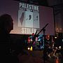A band performs on a dimly lit stage in front of a large screen displaying maps of Palestine from 1947, 1967, and today with the word STOLEN below.