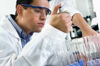 A photo of a man adding chemicals to a series of pipettes, wearing goggles and a lab coat