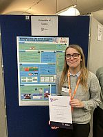 Charlotte Richardson standing in front of her academic poster holding her certificate for attending Posters in Parliament in 2025