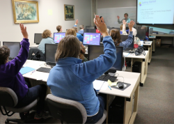 Adults in a classroom setting watching a presentation, some with their hands up