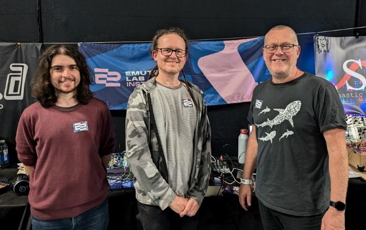 Three founders of Emute Lab Instruments stood smiling in front of their stall at an event
