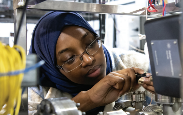 A student leans into a large piece of metal apparatus and attaches parts of the machinery together.