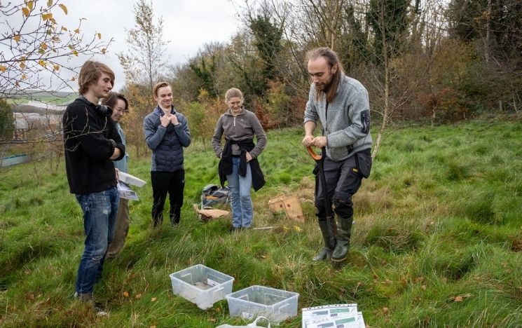 A group of students stand in a green area of campus next to large plastic tubs on the grass