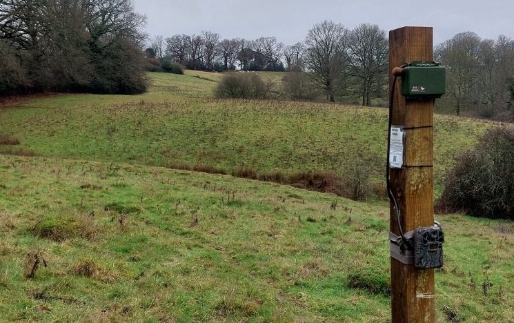 A wooden post with monitoring technology attached to it stands in a field with trees and rolling hills in the distance