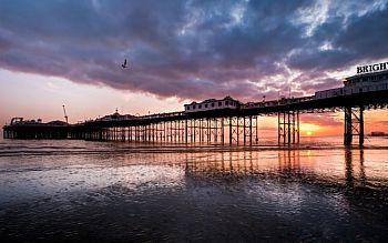 A photo of the pier taken from down on the beach, on the left side of the pier. The tide is very low so there is only sand under the pier, and there is a deep purple, orange sunset.