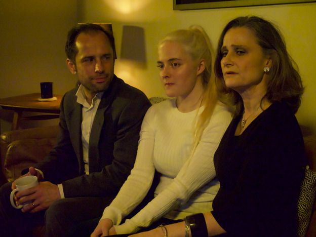Three people (one man, two women) sitting on a settee. The man is looking at the women but the women are looking away. He is holding a white mug.