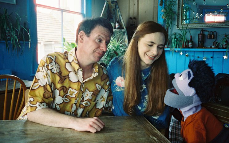 A man and a woman sit at a table looking happily at a male puppet.