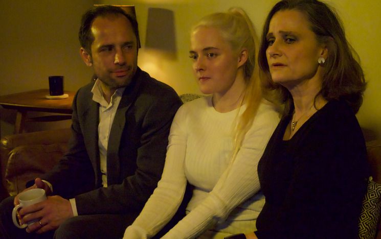 Three people (one man, two women) sitting on a settee. The man is looking at the women but the women are looking away. He is holding a white mug.